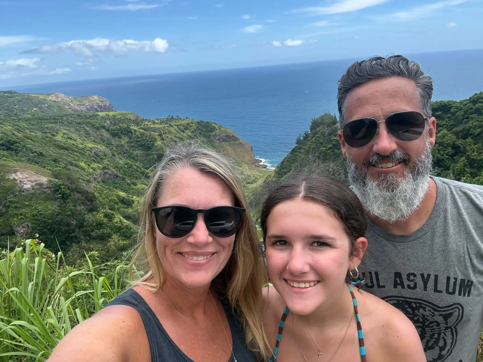 A family of three smiling for a selfie in front of a lush green coastal valley that runs down to the ocean